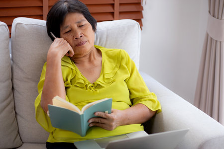 Asian senior woman thoughtful writing a book sitting on sofa with relax in living room at home, elderly woman planning and strategy writing diary, hobby and recreation in weekend.の写真素材