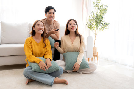 Group young asian friends watching TV together in a cozy living room at home with happy, laughing and enjoying sitting on sofa for watching television with relax, lifestyle and entertainment.の写真素材