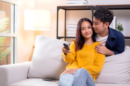 Happy young asian couple sitting on cozy sofa watching TV together and enjoying a relaxation in living room at home, relationship and comfortable of couple during watching television on couch.の写真素材