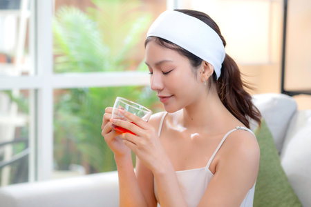 Beautiful young asian woman with headband enjoying tea sitting on sofa with satisfied in living room at home, happy woman drinking tea or beverage for health and relaxation, lifestyle and health.の写真素材