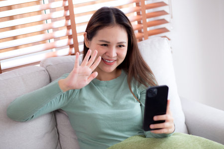 Young asian woman sitting on sofa looking smartphone for video call in living room at home, female using social media on smart phone for chat online and conversation distant, lifestyle concept.の写真素材