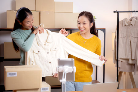 Two young asian woman live online clothing store while showing clothing with tablet on social at home, entrepreneur streaming for selling online clothing and presenting, online seller.の写真素材