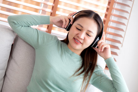 Happy young asian woman wearing headphones listening music while sitting on sofa with fun and enjoy in the living room at home, happiness female with entertainment, one person, lifestyle concept.の写真素材