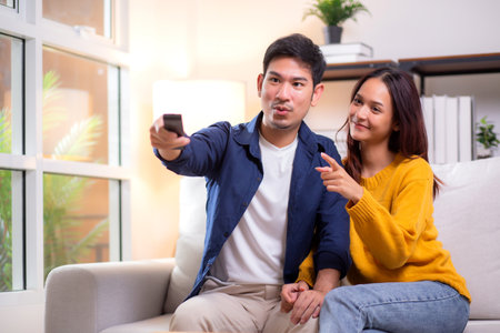 Happy young asian couple sitting on cozy sofa watching TV together and enjoying a relaxation in living room at home, relationship and comfortable of couple during watching television on couch.の写真素材