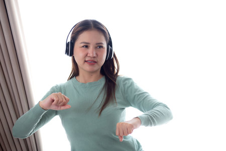 Happy young asian woman wearing headphones listening music while standing dancing with fun and enjoy in the living room at home, happiness female with entertainment, one person, lifestyle concept.の写真素材