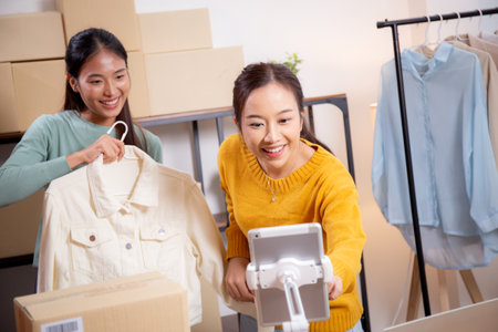 Two young asian woman live online clothing store while showing clothing with tablet on social at home, entrepreneur streaming for selling online clothing and presenting, online seller.の写真素材