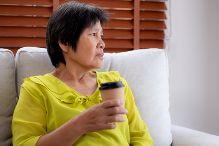 Happiness senior woman sitting on sofa and holding a coffee cup drinking looking out a window in living room at home, elderly drinking beverage for relax, lifestyle and cozy, domestic life.の写真素材