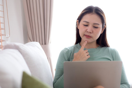 Young asian business woman working with laptop while thoughtful sitting on sofa in home office, businesswoman using on notebook in living room at home, woman thinking idea using laptop.の写真素材