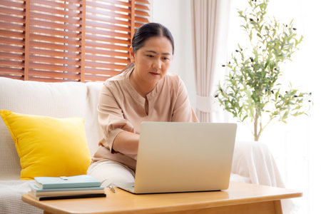 Asian woman working or studying online using a laptop sitting on sofa in living room at home, woman learning for education with determined and inspiration on notebook, e-learning and tutorial.の写真素材