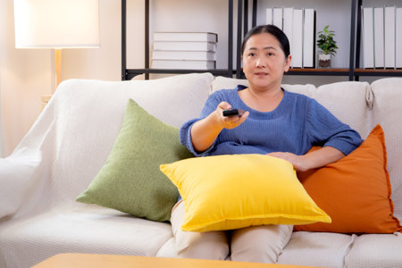 Asian woman sitting on a sofa watching TV with a remote control in living room at home, happy woman relax in a cozy and enjoy entertainment on movie, contentment and lifestyle for fun and satisfied.の写真素材