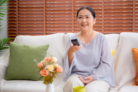 Asian woman sitting on a sofa watching TV with a remote control in living room at home, happy woman relax in a cozy and enjoy entertainment on movie, contentment and lifestyle for fun and satisfied.の写真素材