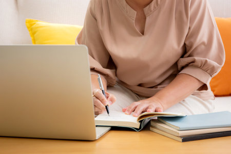 Closeup hands asian woman working or studying online using a laptop and writing on textbook sitting on sofa in living room at home, woman learning for education on notebook, e-learning and tutorial.の写真素材