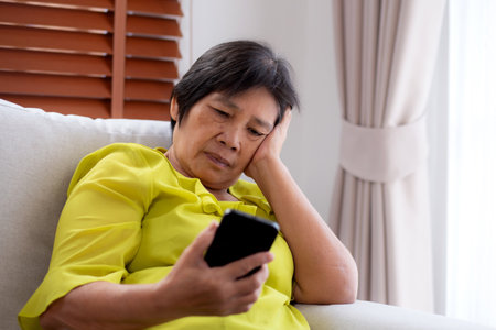 Senior asian woman stress and worried sitting on sofa while looking on smart phone in living room at home, woman unhappy and concerned watching on smartphone, lifestyle and communication.の写真素材