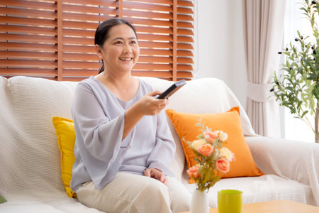 Asian woman sitting on a sofa watching TV with a remote control in living room at home, happy woman relax in a cozy and enjoy entertainment on movie, contentment and lifestyle for fun and satisfied.の写真素材