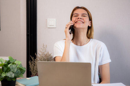 Young caucasian businesswoman talking with smartphone and working on laptop computer in home office, business woman working on laptop and speaking on smart phone, business and communication.の写真素材