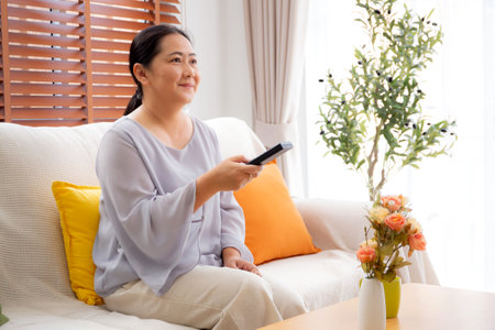 Asian woman sitting on a sofa watching TV with a remote control in living room at home, happy woman relax in a cozy and enjoy entertainment on movie, contentment and lifestyle for fun and satisfied.の写真素材