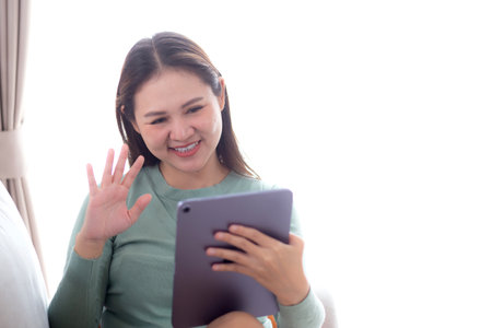 Young asian woman sitting on sofa using digital tablet computer for video call in living room at home, female using social media on tablet for chat online and conversation distant, lifestyle concept.の写真素材