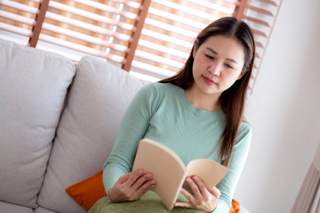 Young asian woman reading a book with expression sitting on sofa in living room at home, woman relax and leisure sitting on couch reading literature, activity and hobby, lifestyle and relaxation.の写真素材