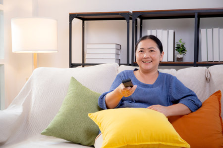 Asian woman sitting on a sofa watching TV with a remote control in living room at home, happy woman relax in a cozy and enjoy entertainment on movie, contentment and lifestyle for fun and satisfied.の写真素材