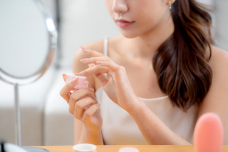 Beauty young asian woman applying cream or lotion while looking mirror in living room at home, happy beautiful woman applying skincare for rejuvenation on facial, skin care and health, self care.の写真素材