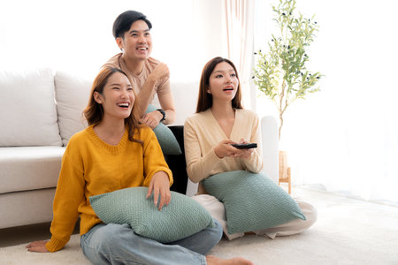 Group young asian friends watching TV together in a cozy living room at home with happy, laughing and enjoying sitting on sofa for watching television with relax, lifestyle and entertainment.の写真素材
