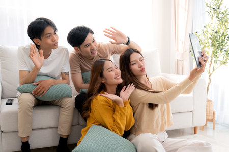 Group young asian friends sitting on sofa taking a selfie or video call using digital tablet in living room at home, cheerful and smiling while connection to social media, interaction and friendship.の写真素材