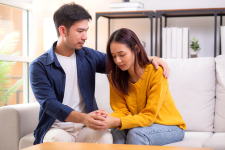 Young asian caring man comforts sad woman with gentle embrace sitting on sofa in living room at home, wife with expression unhappy and anxious while husband consoling with tenderness and closeness.の写真素材