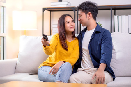 Happy young asian couple sitting on cozy sofa watching TV together and enjoying a relaxation in living room at home, relationship and comfortable of couple during watching television on couch.の写真素材