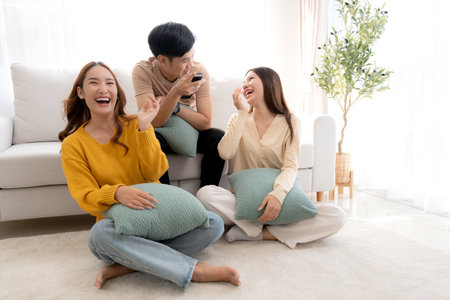 Group young asian friends watching TV together in a cozy living room at home with happy, laughing and enjoying sitting on sofa for watching television with relax, lifestyle and entertainment.の写真素材