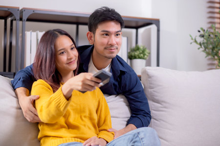 Happy young asian couple sitting on cozy sofa watching TV together and enjoying a relaxation in living room at home, relationship and comfortable of couple during watching television on couch.の写真素材