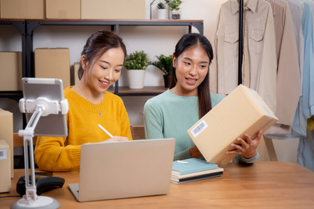 Two young asian woman working while planning online business and checking order with writing on tablet at home, retail with online seller, startup business and cargo, shipping and service.の写真素材