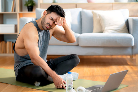 Tired young asian man sitting on a yoga mat after a home workout wiping sweat in living room at home, exhausted young man after exercise watching laptop in cozy, self-care and wellbeing.の写真素材