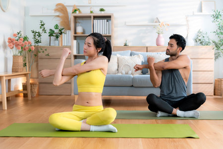 Young asian couple practicing yoga in living room at home, stretching arm pose, fitness and flexibility on yoga mat, man and woman doing yoga together in a cozy home, self-care for health.の写真素材
