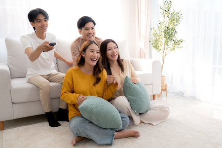 Group young asian friends watching TV together in a cozy living room at home with happy, laughing and enjoying sitting on sofa for watching television with relax, lifestyle and entertainment.の写真素材