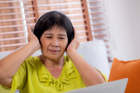 Happy senior asian woman wearing headphones listening music while sitting on sofa with fun and enjoy in the living room at home, happiness elderly female with entertainment, lifestyle and activities.の写真素材