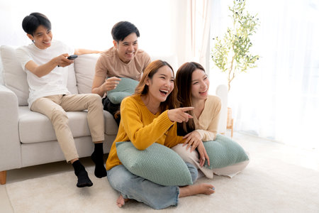Group young asian friends watching TV together in a cozy living room at home with happy, laughing and enjoying sitting on sofa for watching television with relax, lifestyle and entertainment.の写真素材
