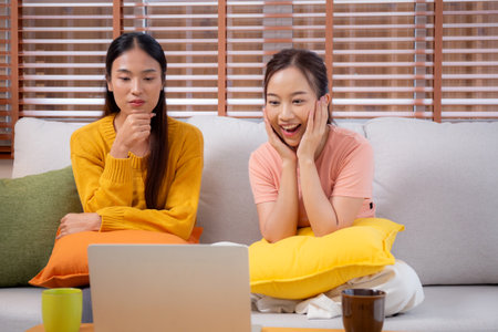 Two young asian woman sitting on sofa using laptop computer for video call in living room at home, female using social media on notebook for online and conversation distant, lifestyles concept.の写真素材