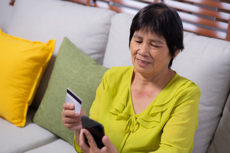 Senior asian woman sitting on sofa using smart phone shopping online with credit card buying to internet, female payment with business with smartphone, purchase and payment, business and finance.の写真素材