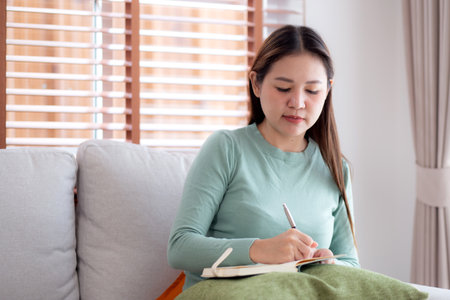 Happy young asian woman writing on book sitting on sofa with relax in living room at home, woman studying and education writing on textbook sitting on couch with leisure, lifestyle and hobby.の写真素材