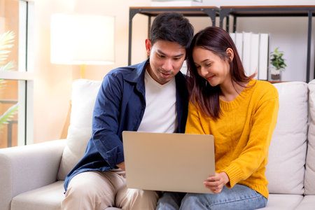 Young asian couple looking on laptop for relax with cozy and contentment sitting on sofa in living room at home, couple leisure watching laptop on couch, relationship and bonding, lifestyle concept.の写真素材