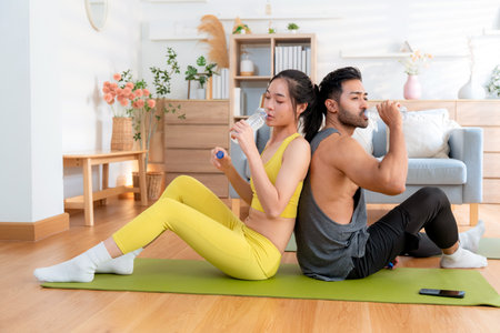 Young asian couple resting together after home workout in living room at home, sitting back to back on yoga mat while drinking water after exercise, refreshment and wellbeing, taking a break.の写真素材