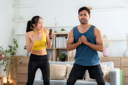 Young asian couple workout during woman hold smartphone encouragement to partner while man doing squat with motivation, woman cheering man with exercise with strong and strength, self-care and health.の写真素材