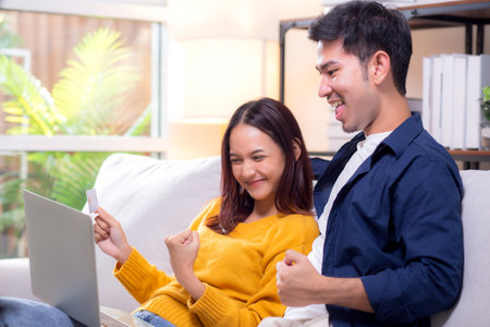 Happy asian couple using laptop and credit card for online shopping together in living room at home, man and woman smiling and relax with enjoy a digital lifestyle, convenient payment.の写真素材