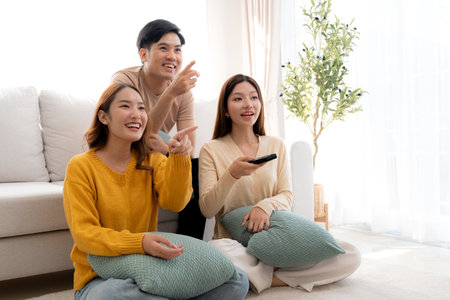 Group young asian friends watching TV together in a cozy living room at home with happy, laughing and enjoying sitting on sofa for watching television with relax, lifestyle and entertainment.の写真素材