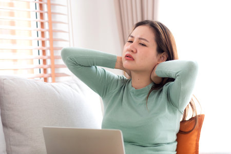 Asian woman sitting on sofa with neck pain while using laptop computer in living room at home, woman sitting on couch using notebook while pain muscle and inflammation with neck, medical concept.の写真素材