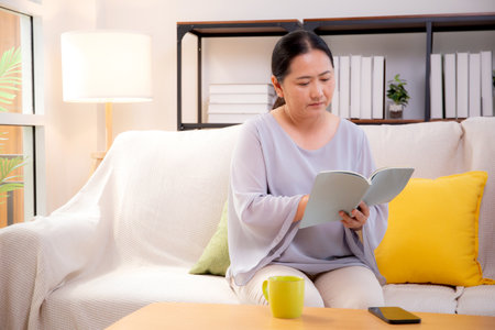 Asian senior woman reading a book sitting on sofa with relax in living room at home, elderly woman reading literature on couch with cozy, hobby and recreation in weekend, lifestyles concept.の写真素材