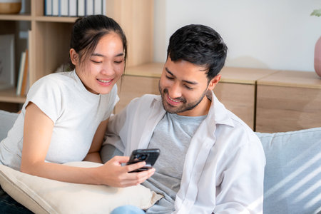 Young asian couple relax sitting on sofa looking smartphone together in living room at home, cozy lifestyle and bonding, relationship and leisure, sharing and enjoy digital content with happy.の写真素材