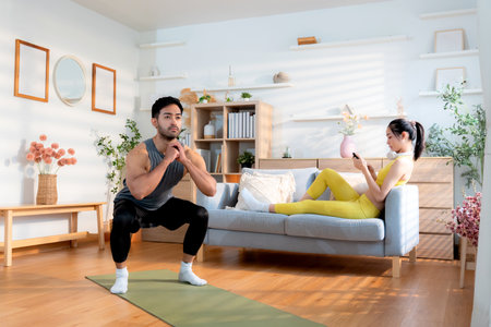 Young asian man doing squat exercise on yoga mat in living room at home while woman using phone for relax on sofa, fitness training and healthy lifestyle in a cozy, strength and strong of muscle.の写真素材