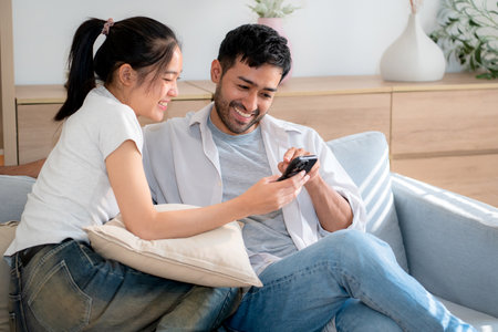 Young asian couple relax sitting on sofa looking smartphone together in living room at home, cozy lifestyle and bonding, relationship and leisure, sharing and enjoy digital content with happy.の写真素材