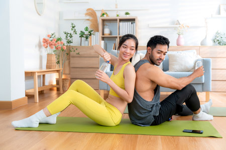 Young asian couple resting together after home workout in living room at home, sitting back to back on yoga mat while drinking water after exercise, refreshment and wellbeing, taking a break.の写真素材